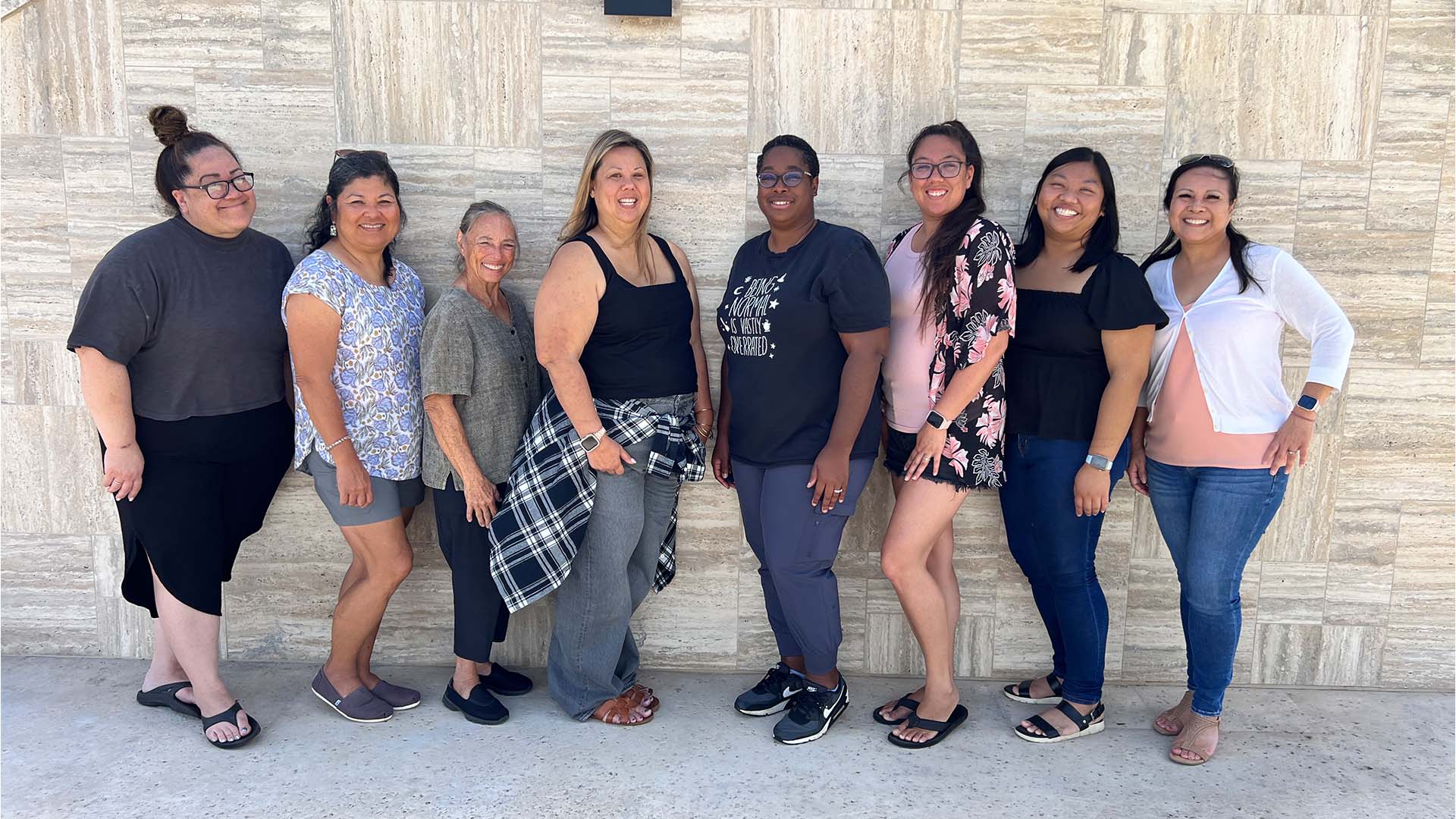 A group of eight women standing in a line, smiling, and posing in front of a textured wall.