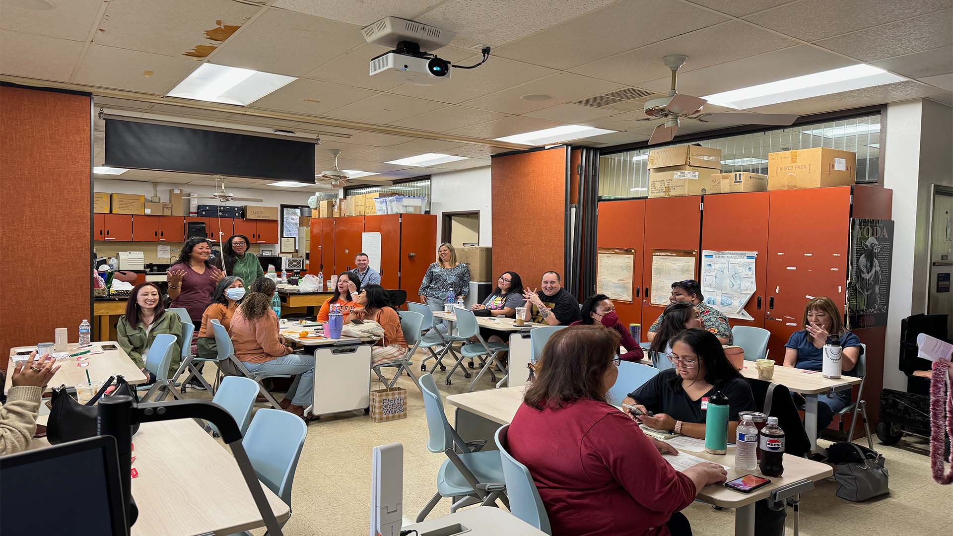 A group of people seated at tables in a classroom setting, engaging in a workshop or meeting, with some participants standing and interacting in the background.